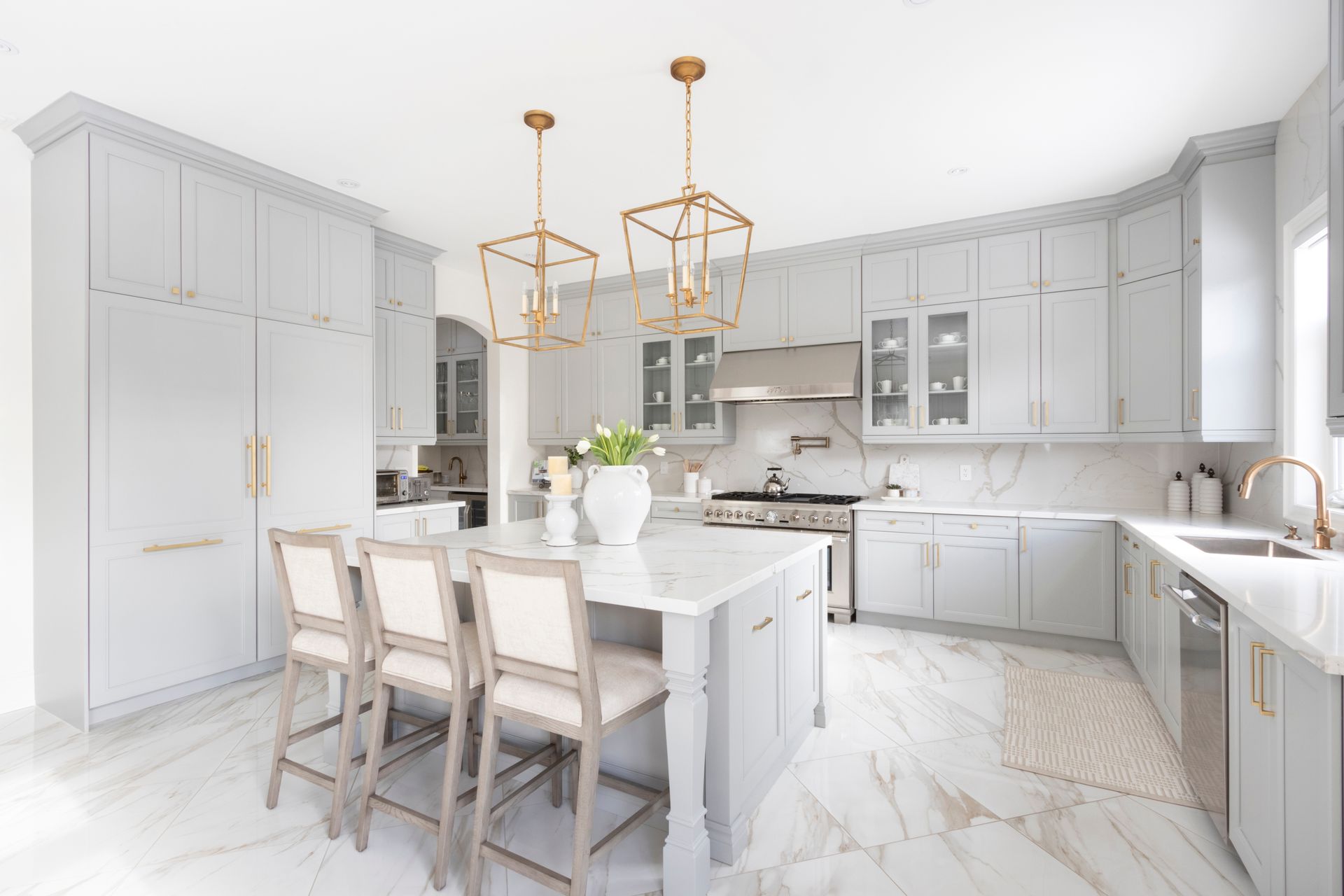 A bright, modern kitchen with light gray cabinetry, a marble-topped island with three bar stools, and two gold pendants.
