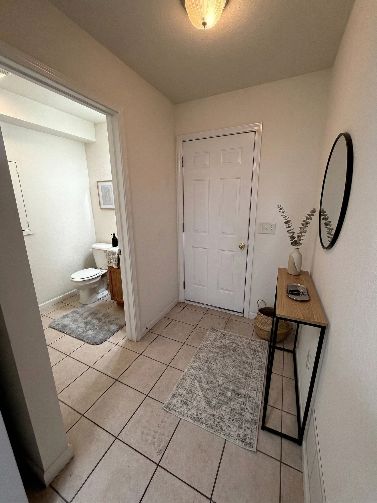 Small tiled hallway with a white door, mirror, and narrow table; bathroom visible through open doorway.