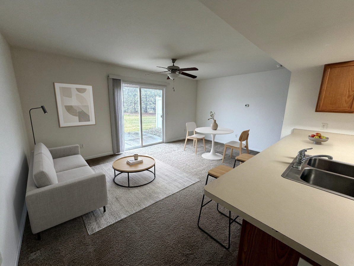 Bright living room with gray sofa, round coffee table, dining nook, ceiling fan, and kitchen counter.