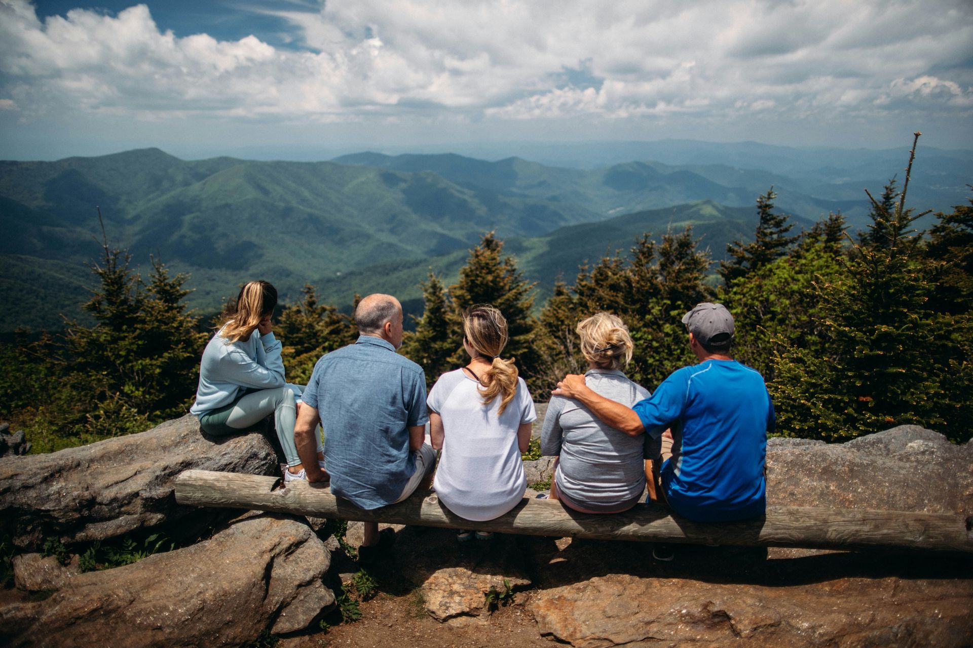 Five hikers sit on a rock ledge overlooking green mountains under a cloudy sky.