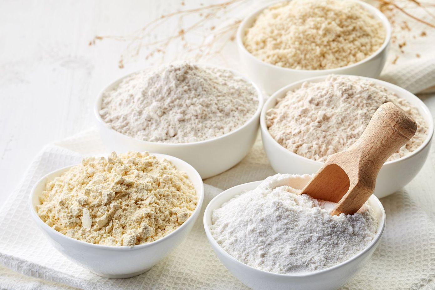 Diverse ciotole contenenti farina e cereali, con mestoli di legno, su un tavolo bianco.