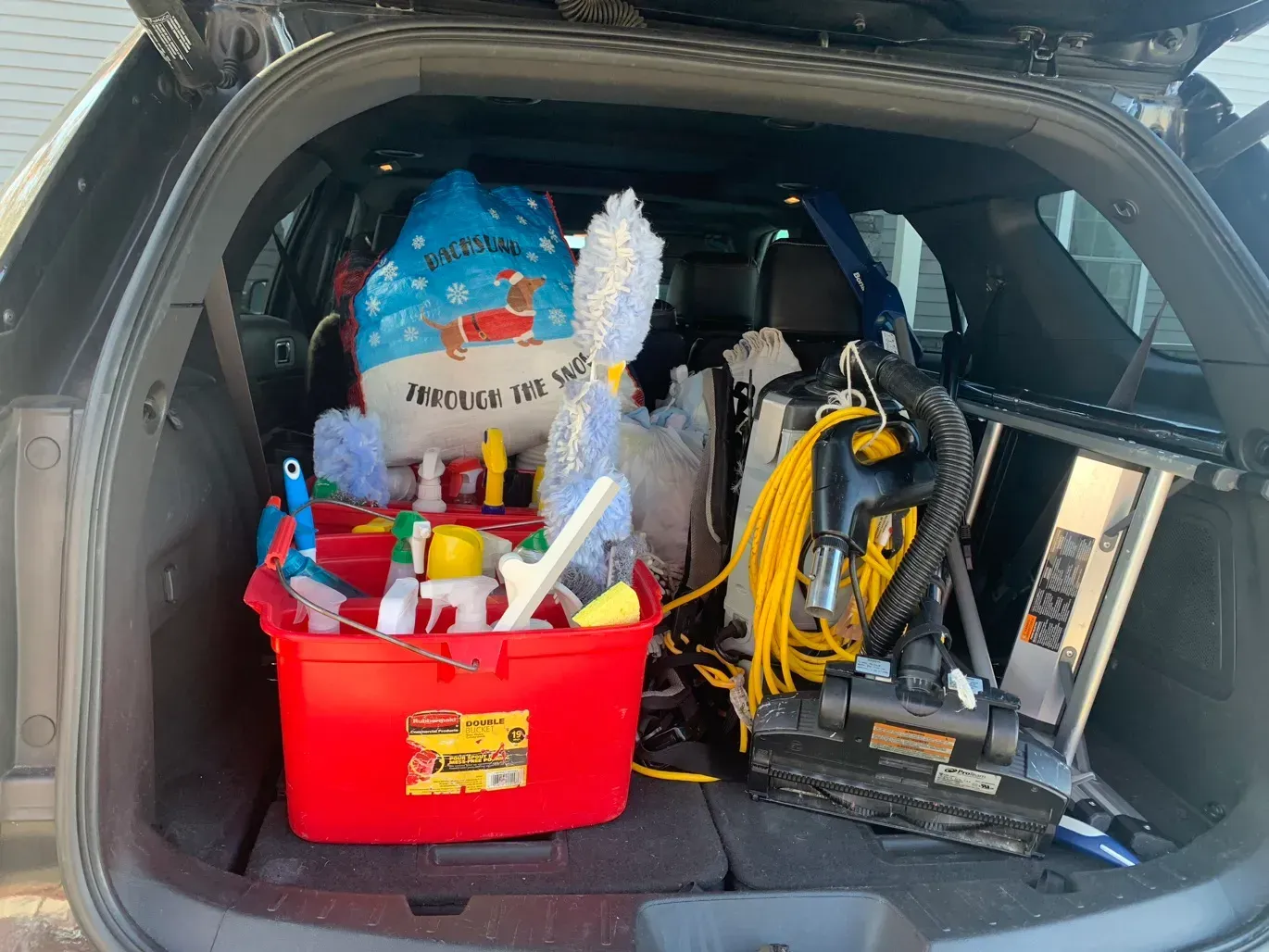 The back of a car is filled with cleaning supplies and a vacuum cleaner.