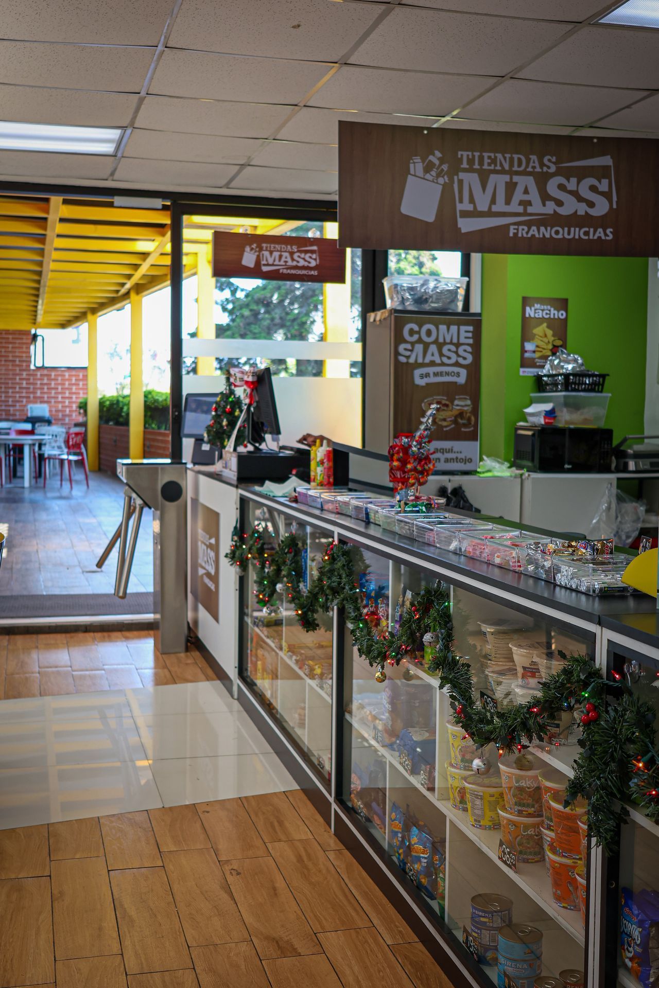 Interior de una tienda de conveniencia con mesas, estantes de productos y una persona detrás del mostrador.