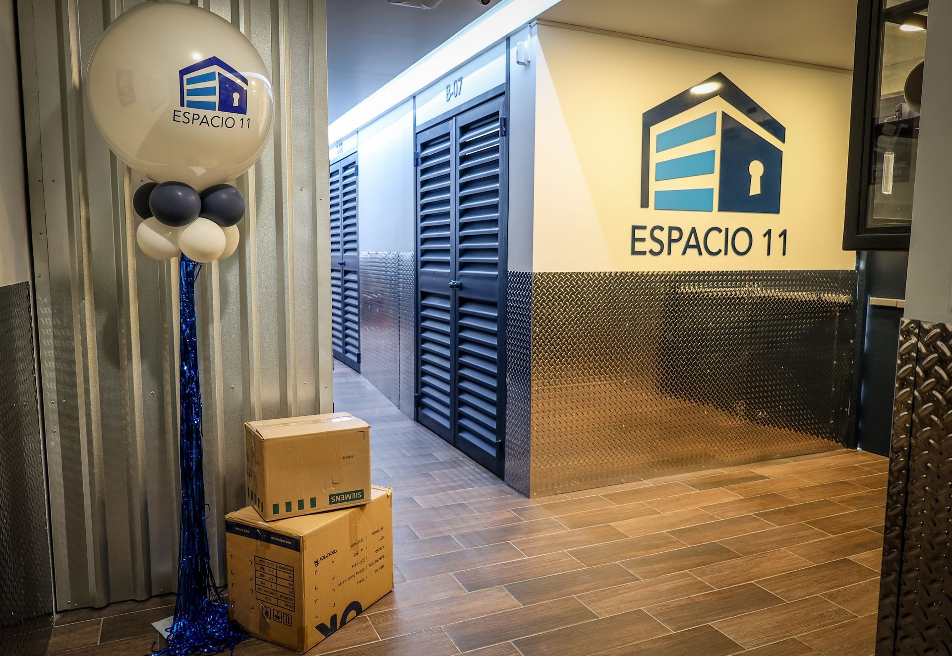 Pasillo del almacén con cajas apiladas y un expositor de globos. Logotipo de Espacio 11 en la pared.