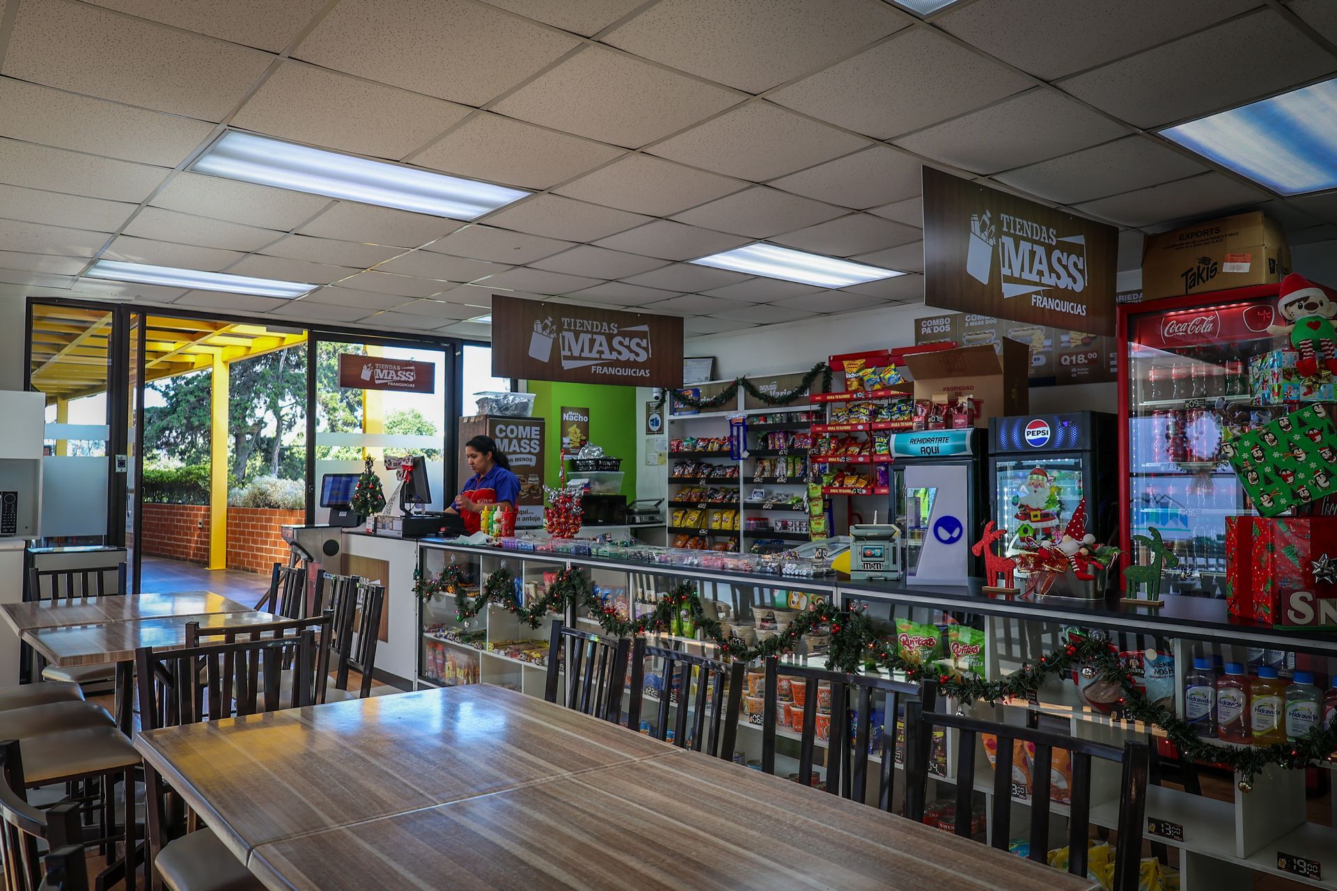 Interior de una tienda de conveniencia con mesas, estantes de productos y una persona detrás del mostrador.