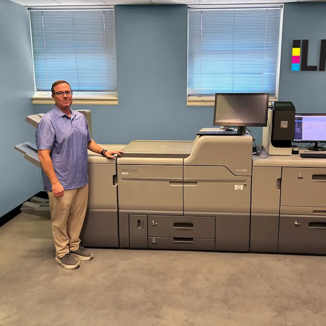 Man standing next to a large printing press in an office; blue walls, windows, LPi logo.