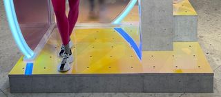 Person in pink leggings and white sneakers steps up neon-lit, yellow-and-blue steps.