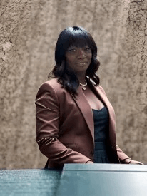 A woman in a brown jacket is standing in front of a wall.