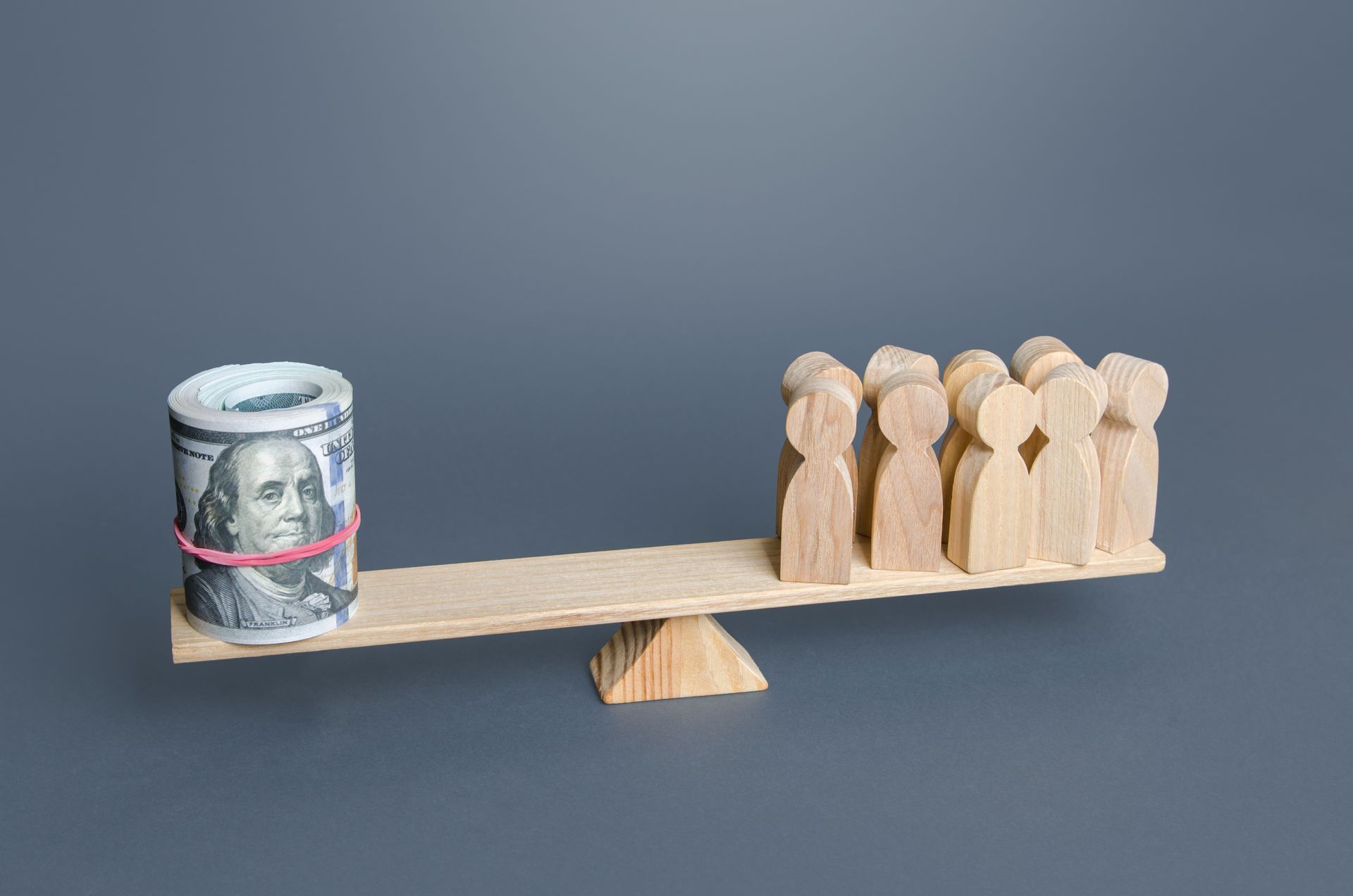 A seesaw balances a roll of cash against a group of wooden figures, symbolizing wealth disparity.