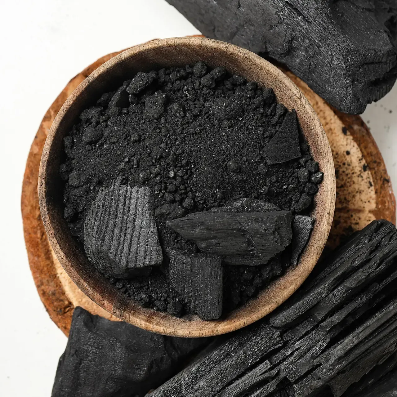 Charcoal pieces, including powder, in a wooden bowl. Black and brown tones on white surface.