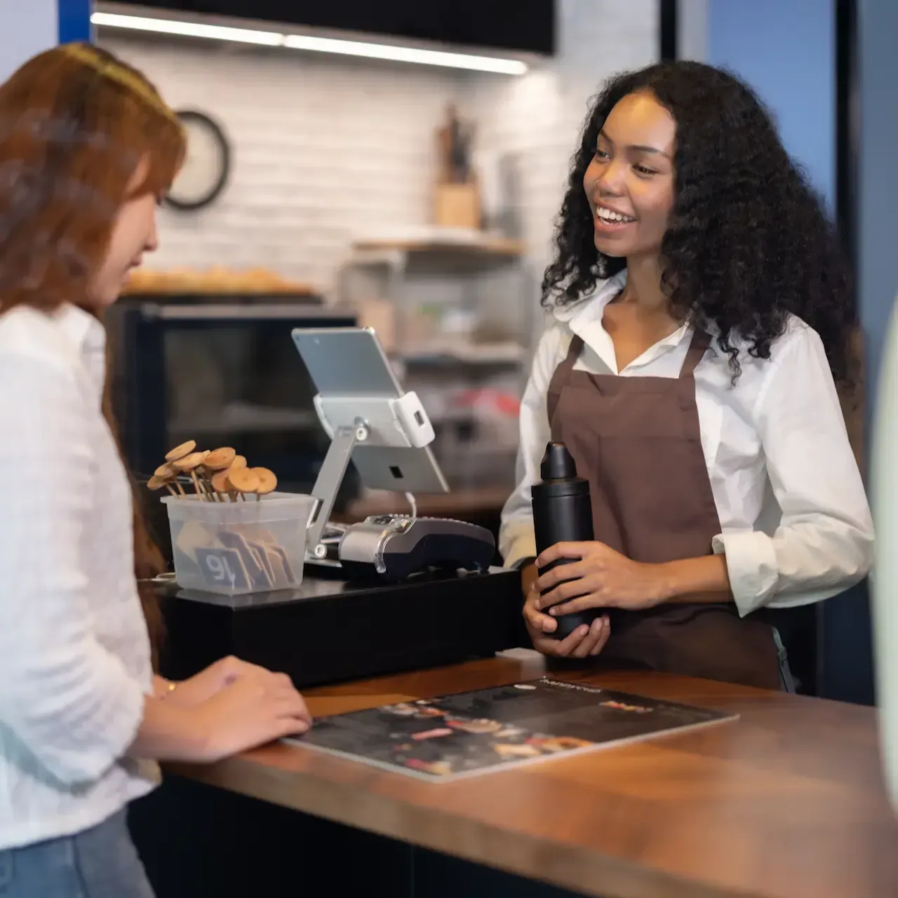 A barista in a brown apron smiles at a customer at a cafe counter.