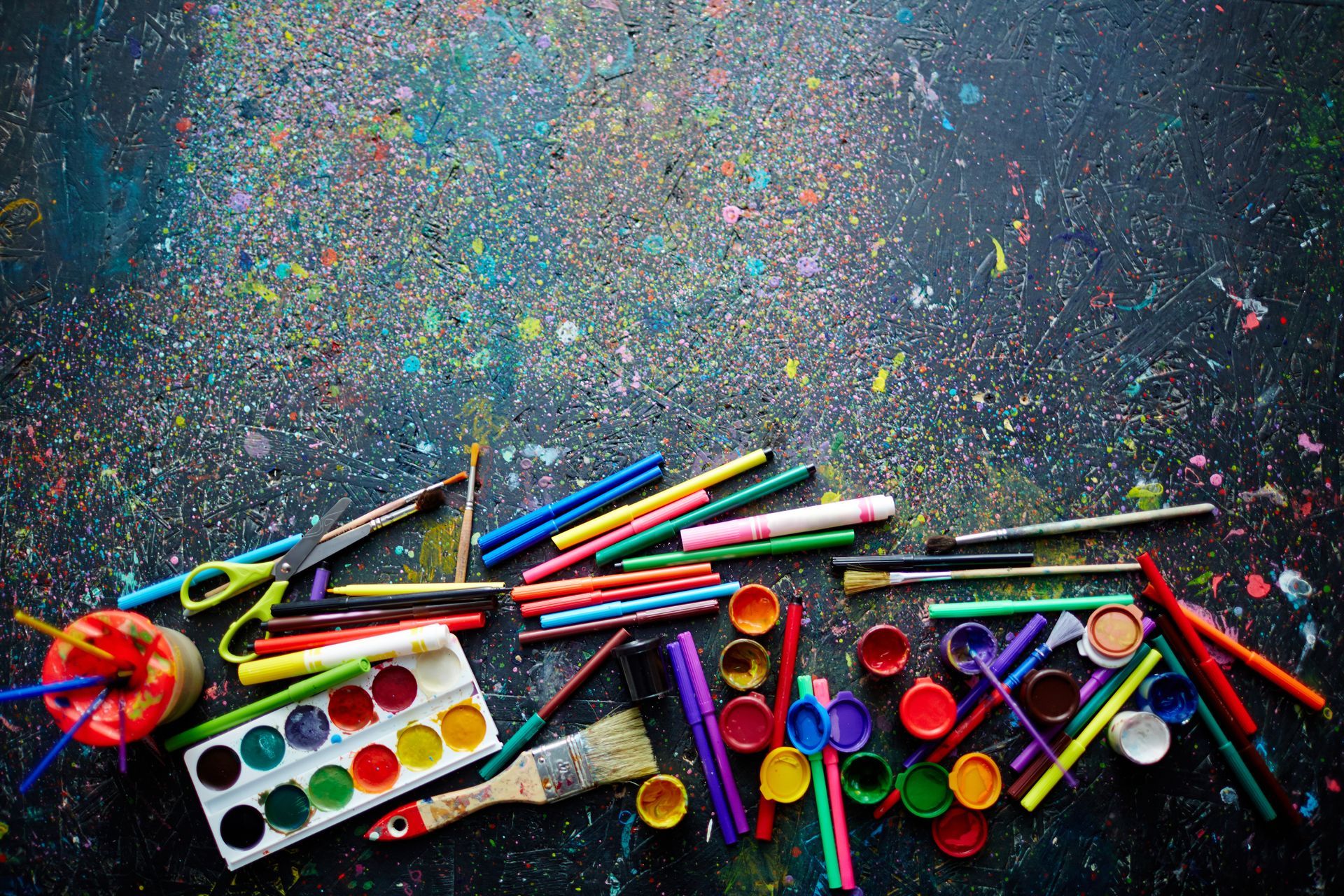 Art supplies scattered on a paint-splattered black surface, including pens, brushes, and watercolor paints.