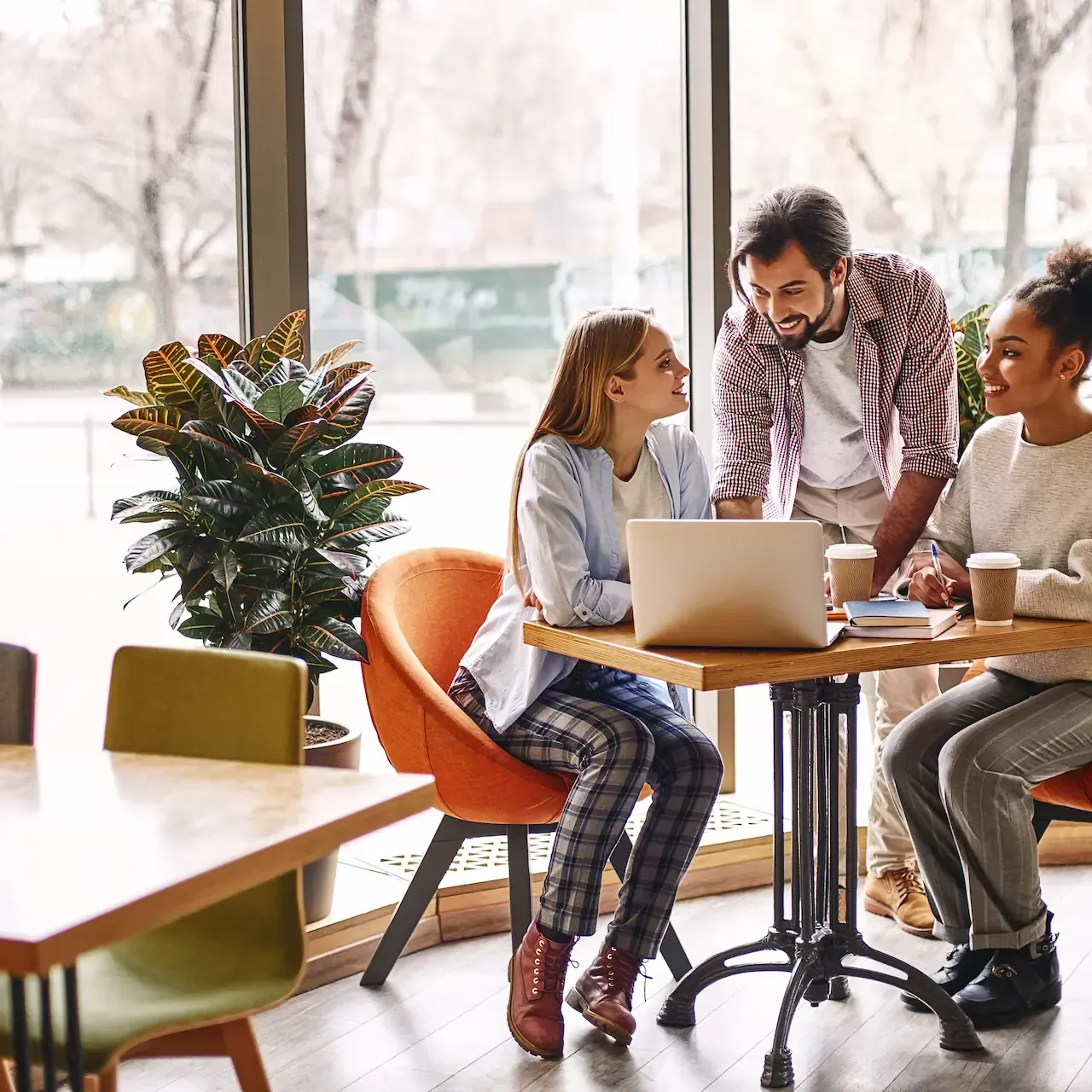 Three people collaborating around a laptop at a table in a cafe, with plants and a large window.