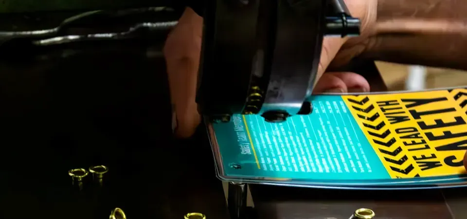 A hand operates a tool, puncturing a teal-colored surface with a safety slogan.