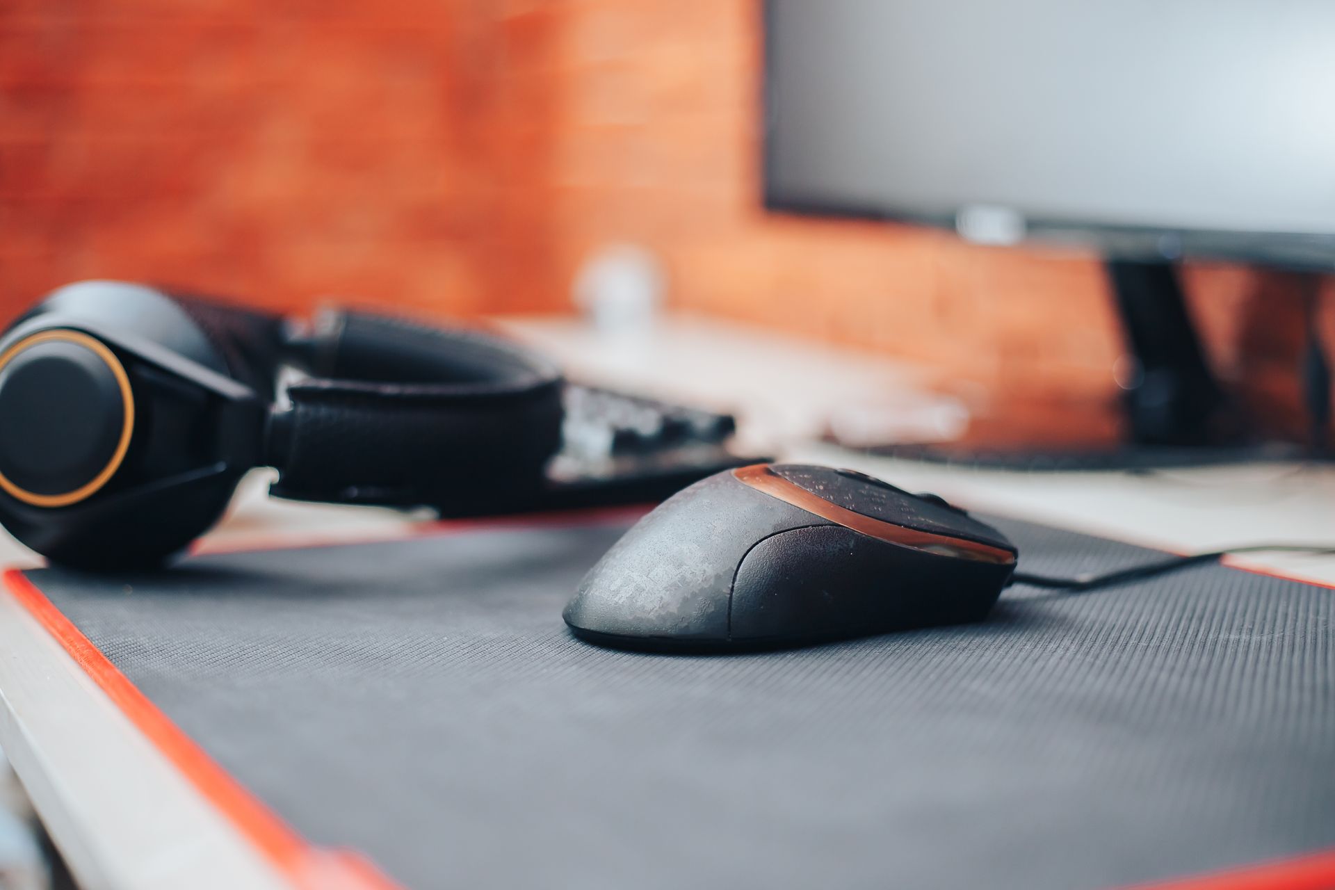 Black computer mouse on mousepad next to black headphones, in front of computer monitor, brick wall background.