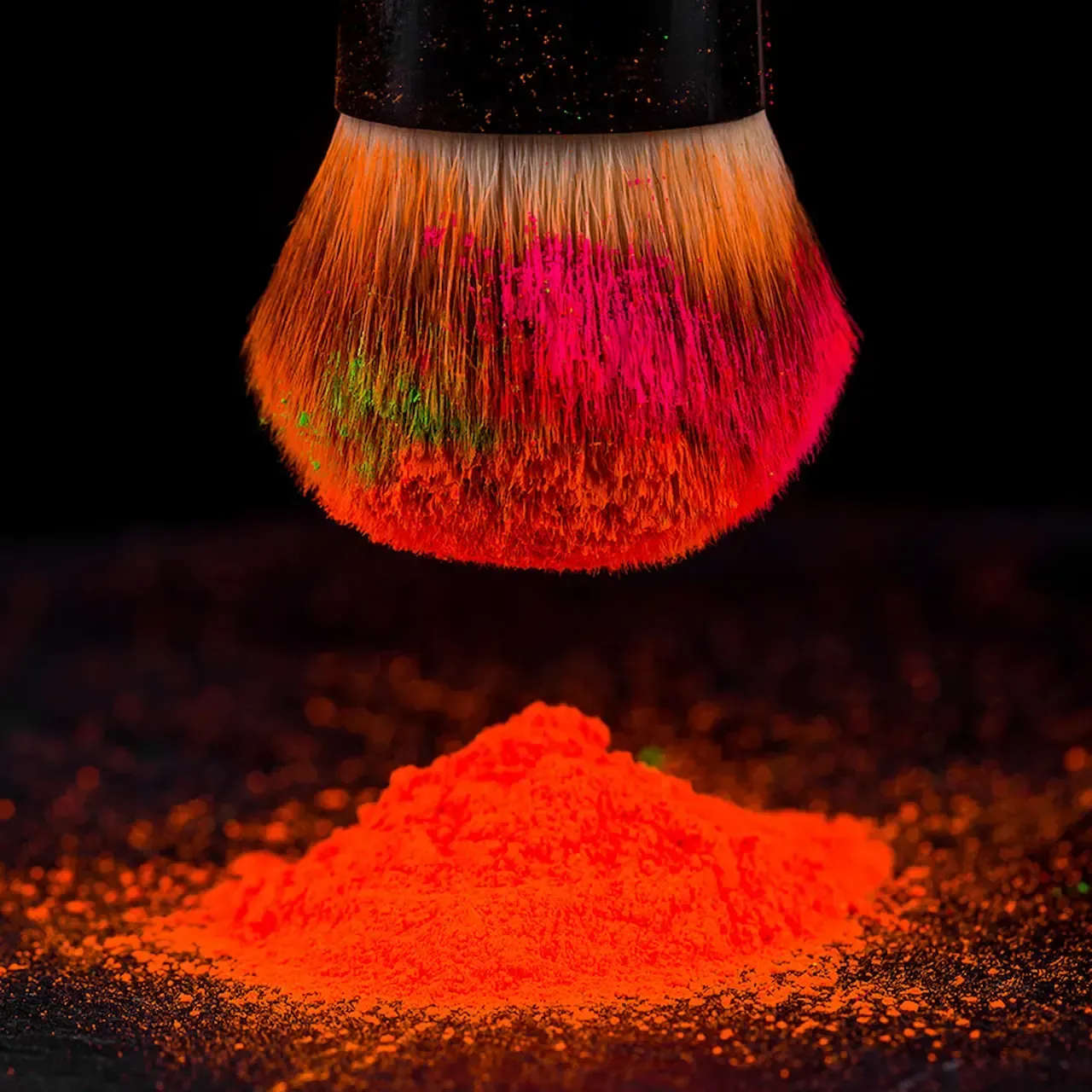 Makeup brush above pile of neon orange powder, with hints of pink and green, against black.
