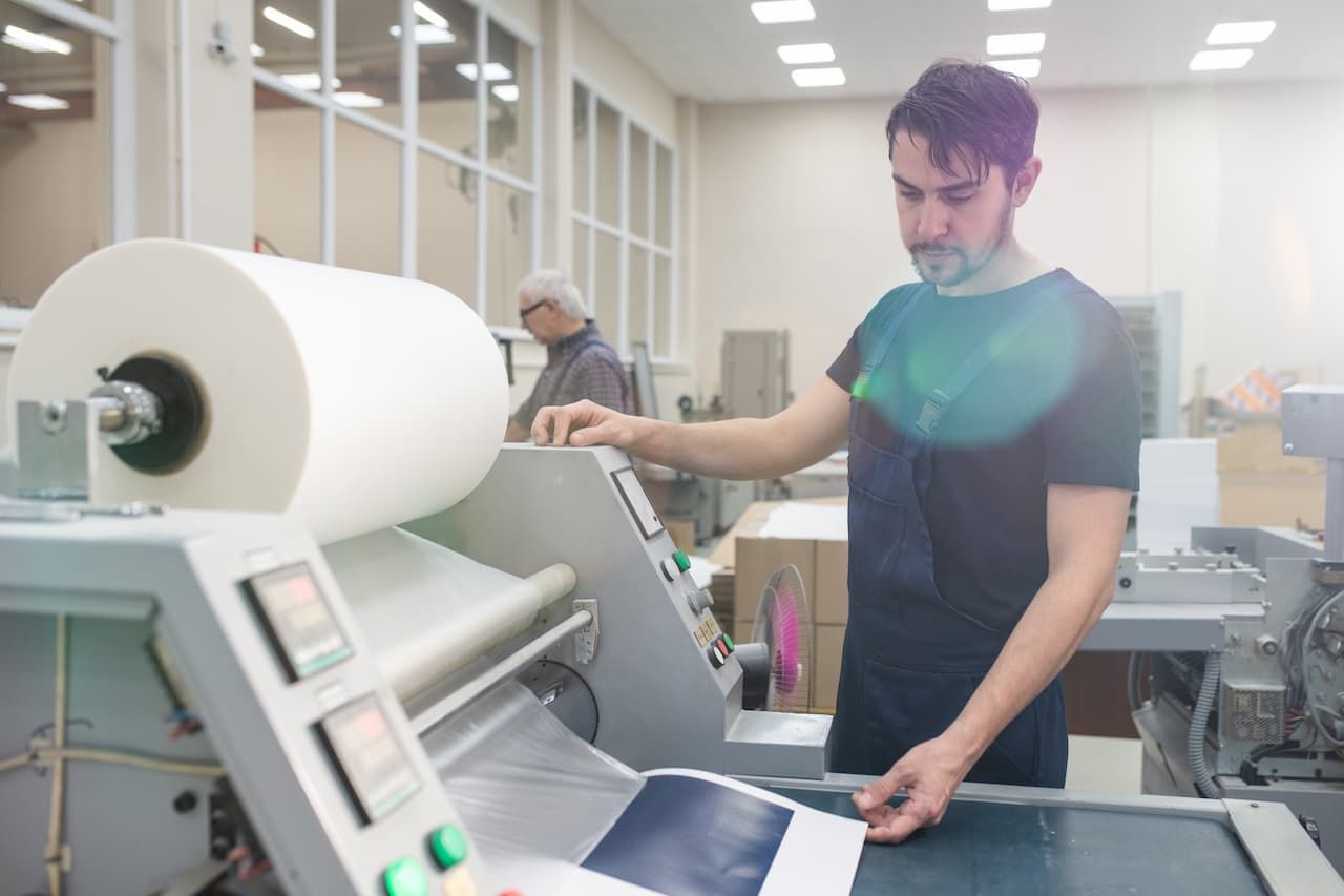 Man operating printing press, inspecting a printed sheet. Another person in the background, rolls of paper.