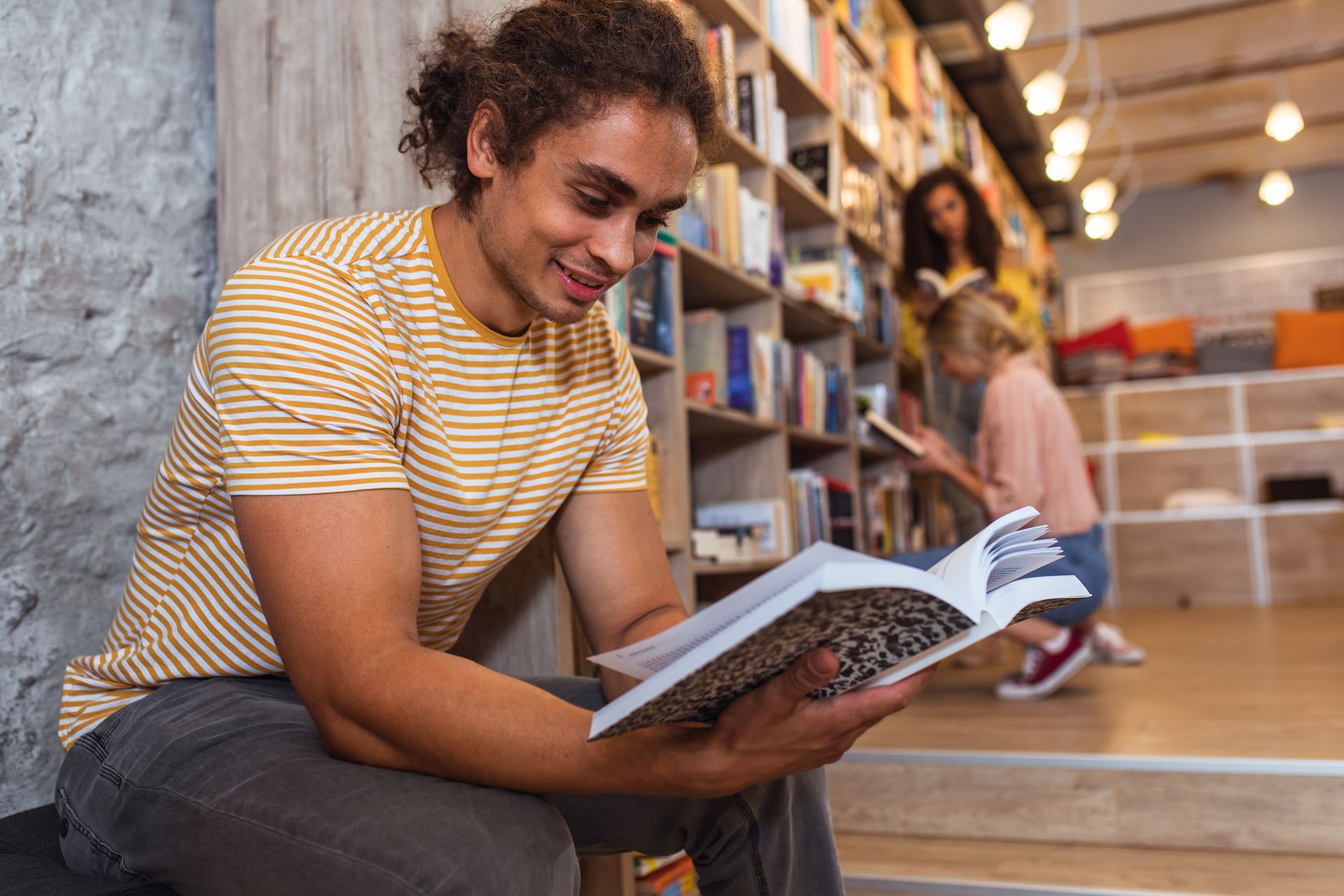 Man reading a book in a library, smiling. Other people browsing in the background.