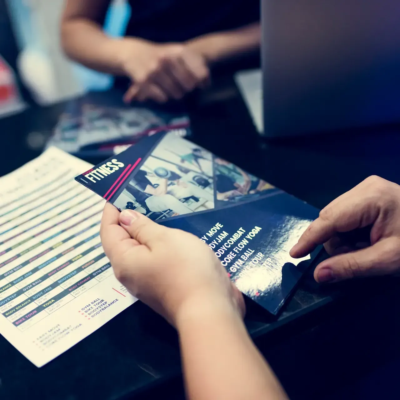 Hands holding a brochure at a desk with papers and a laptop; person behind the desk.