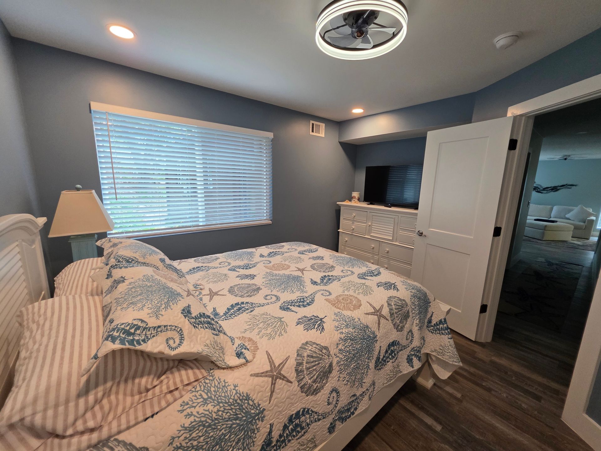 Bedroom with blue walls, beach-themed bedding, dresser with TV, and open door to another room.