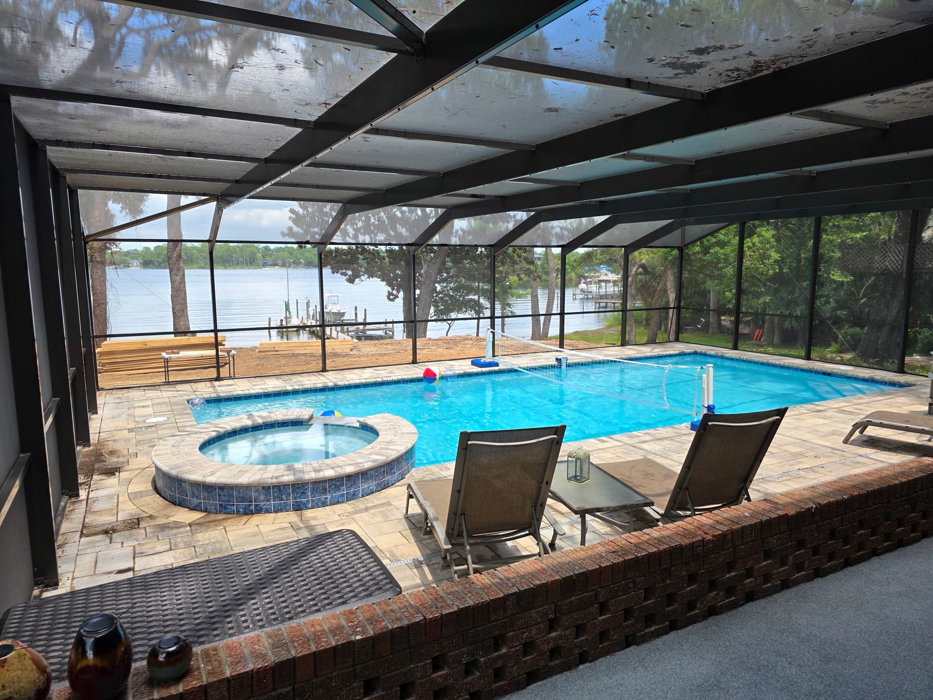 Enclosed pool and hot tub overlooking a bay. Two chairs, blue water, and a screen enclosure.