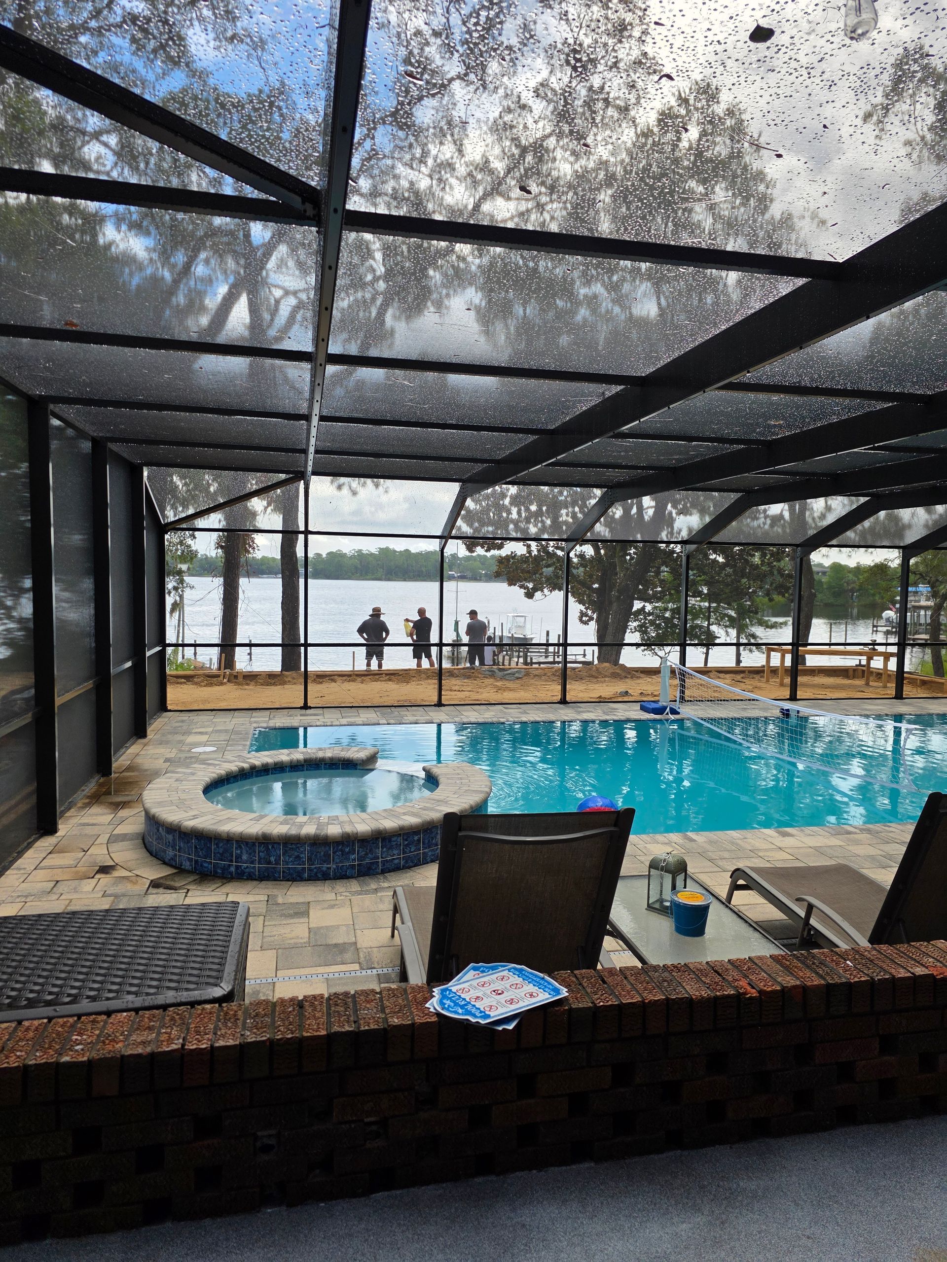 Pool and hot tub under a screened enclosure, overlooking a lake with people on the shore.