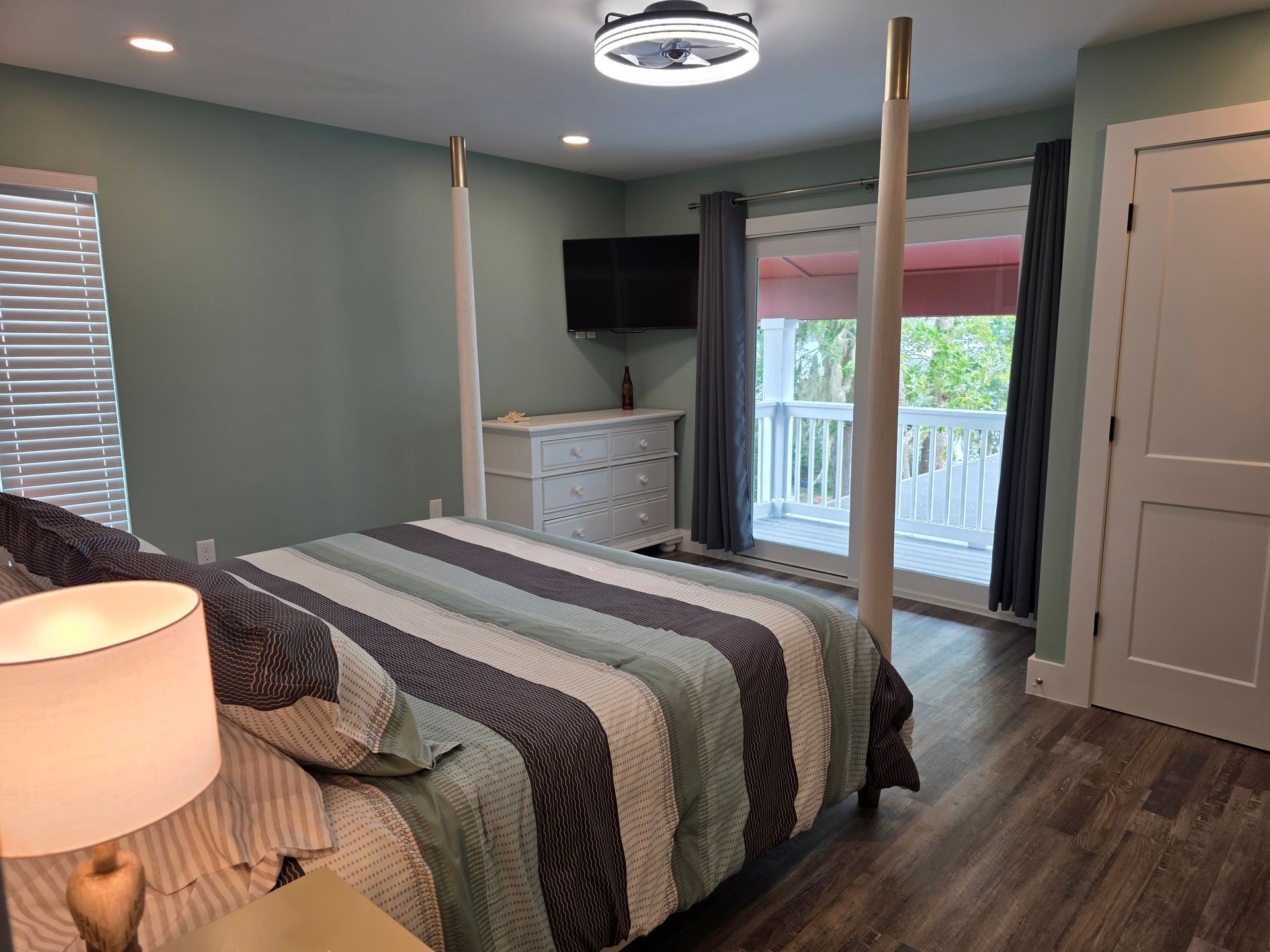 Bedroom with four-poster bed, TV, dresser, and a balcony. Soft green walls, blue and white bedding, light-toned wood floors.