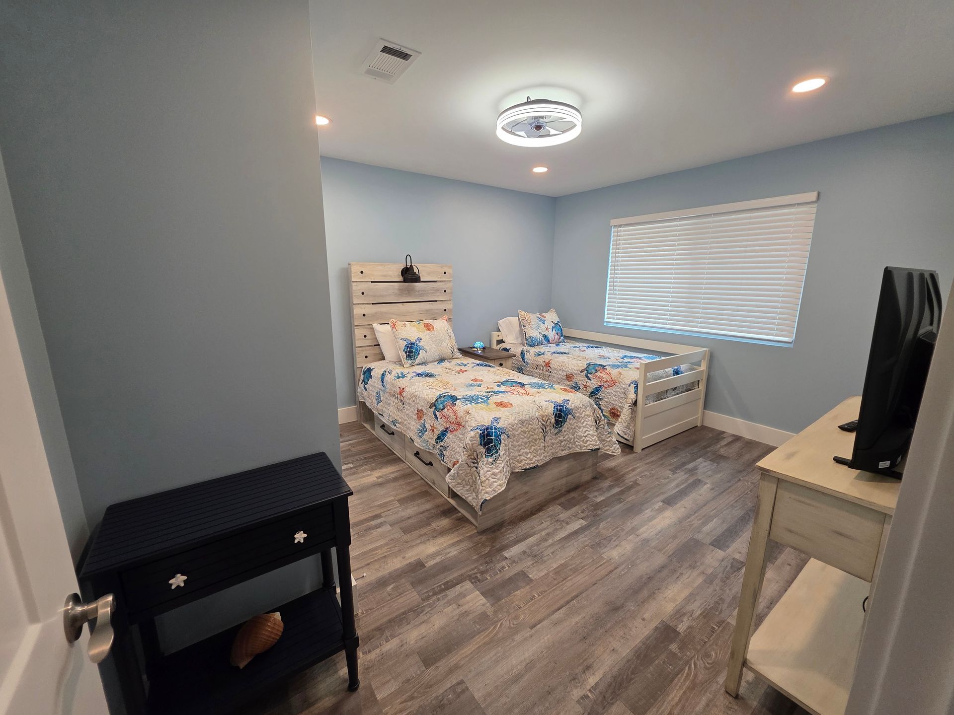 A bedroom with two twin beds, blue walls, and wood-look flooring. Includes a nightstand and a TV.