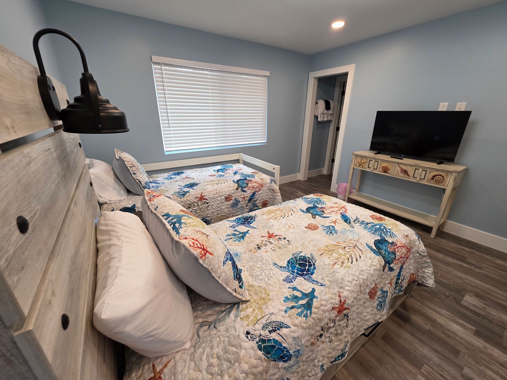 Bedroom with two beds, ocean-themed quilt, TV on a console, window with blinds. Blue walls, wood-look floor.