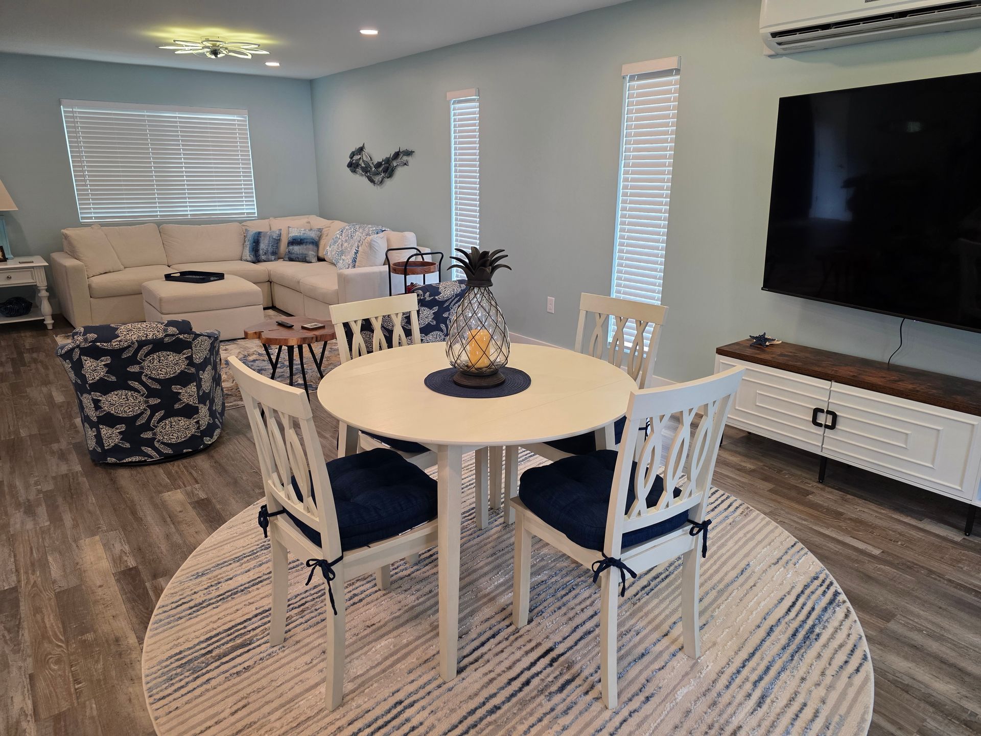 Living room with blue walls, white furniture, round dining table with blue cushions, and a large TV.