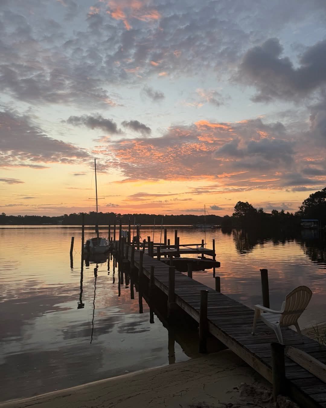 Sunset over a lake with a wooden dock and sailboat. Soft pinks and oranges in the sky reflected in the water.