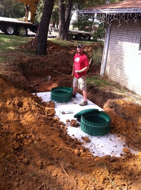 A man in a red shirt is standing in the dirt near a septic tank