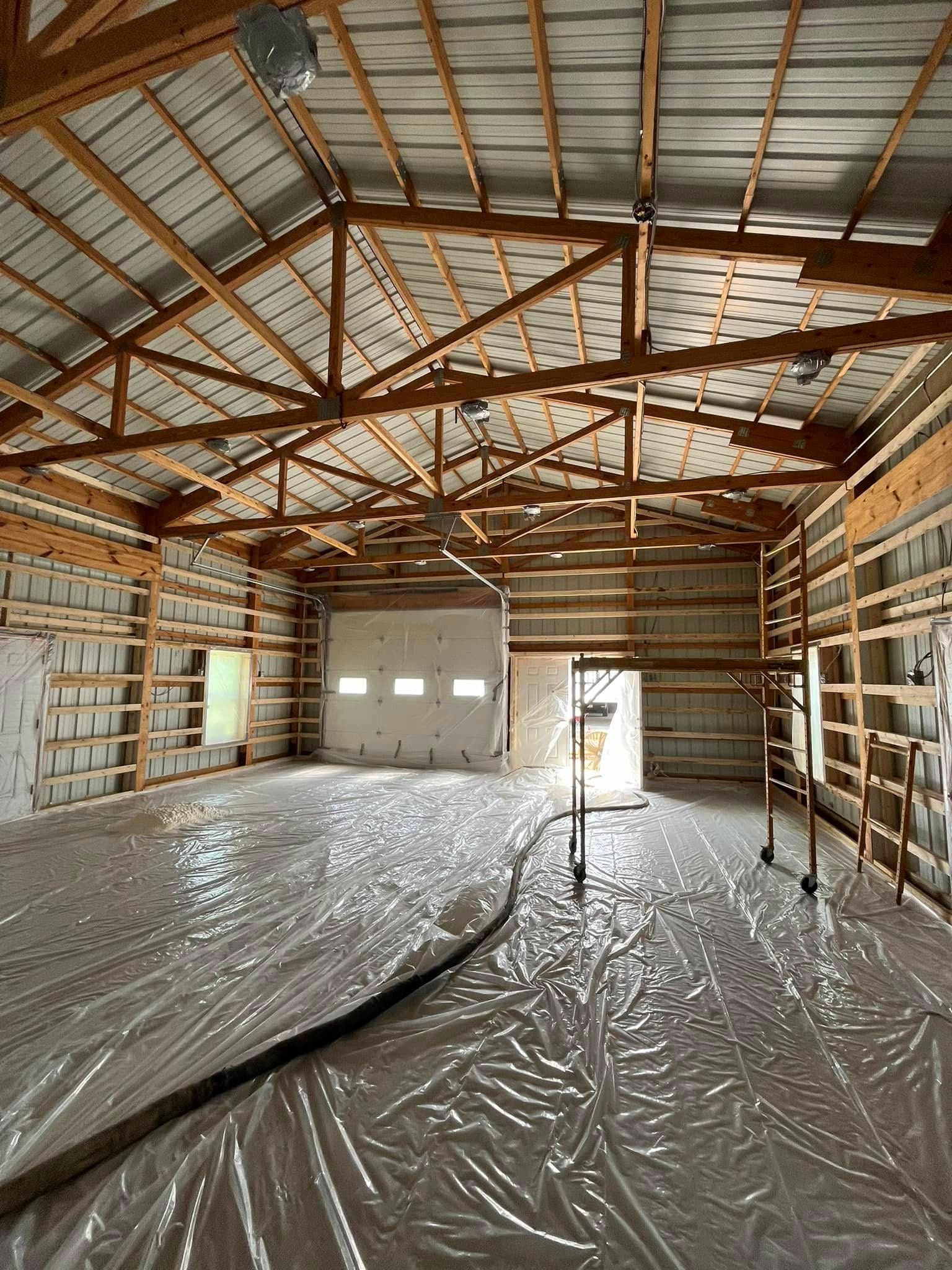 Interior of a large unfinished building with wooden framing, corrugated metal roof, and plastic sheeting on the floor.