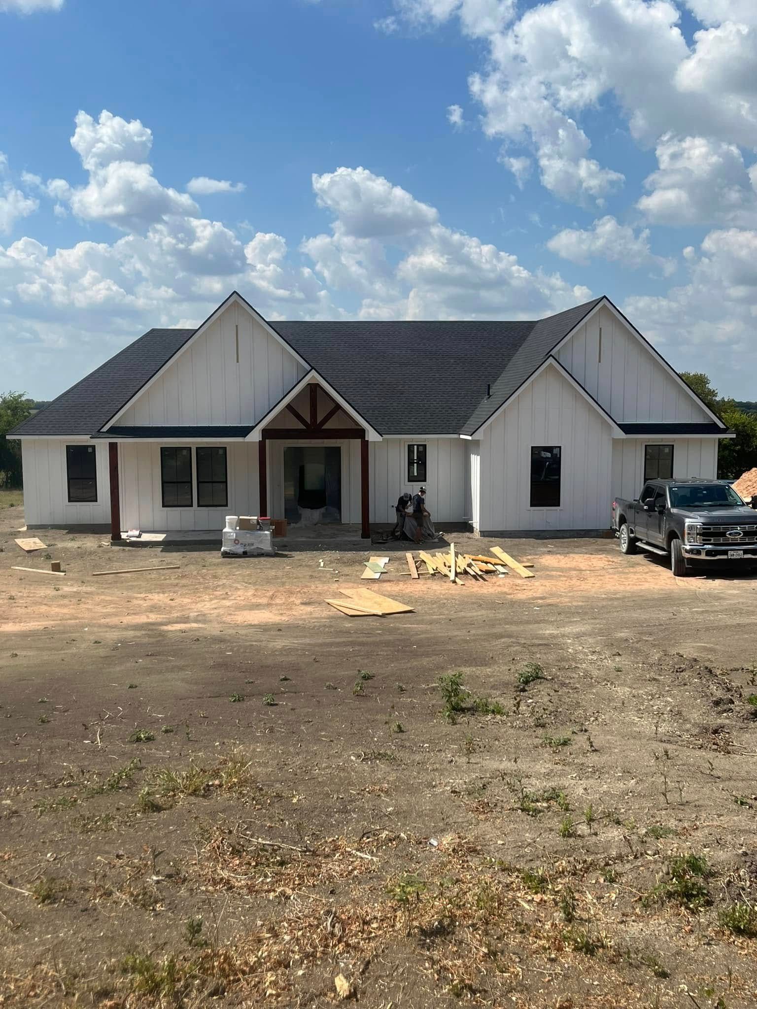 New white farmhouse under construction, dark roof, brown trim, truck parked beside it.