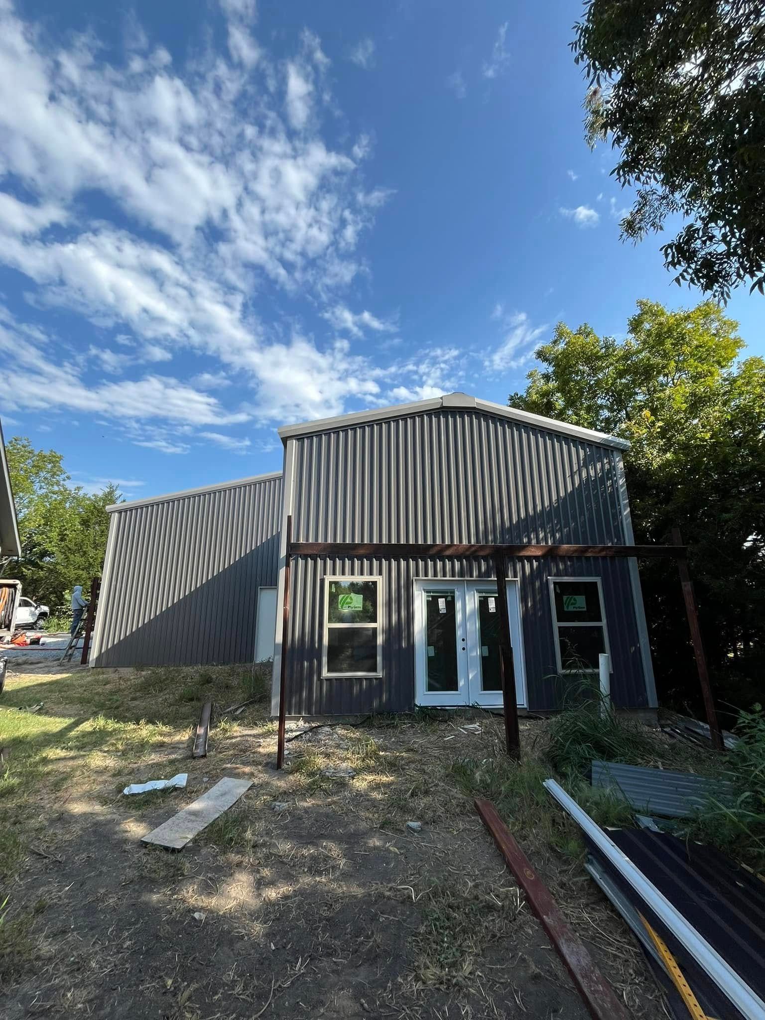 Metal-clad building under construction; corrugated exterior, windows, doorway. Brown metal supports in front, blue sky.