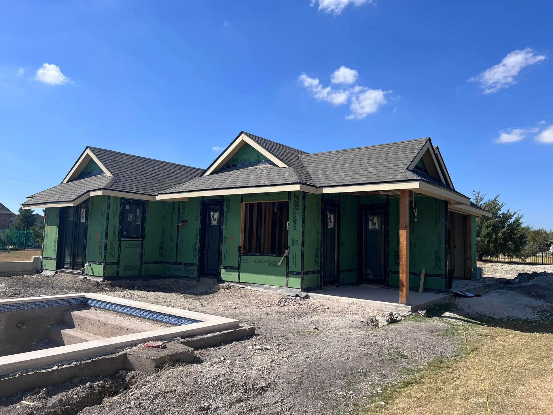 House under construction with green siding, gray roof, and unfinished pool. Blue sky.