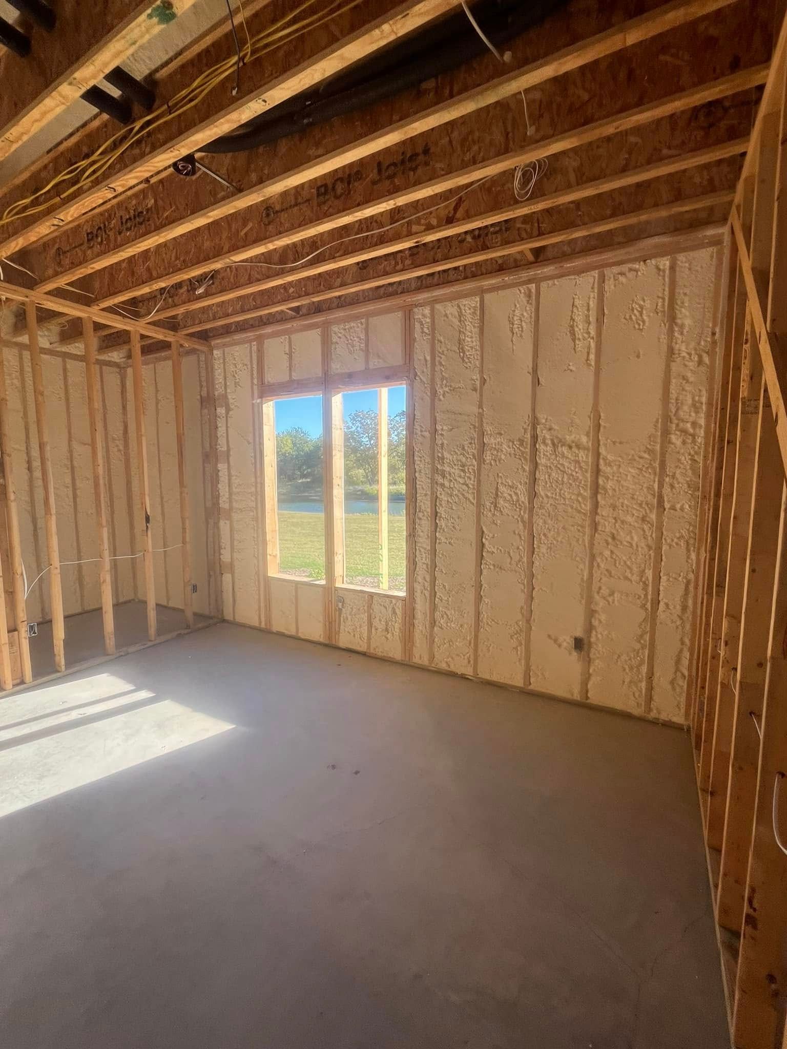 Interior view of a room under construction with exposed wooden framing and spray foam insulation. A window reveals an outdoor view.