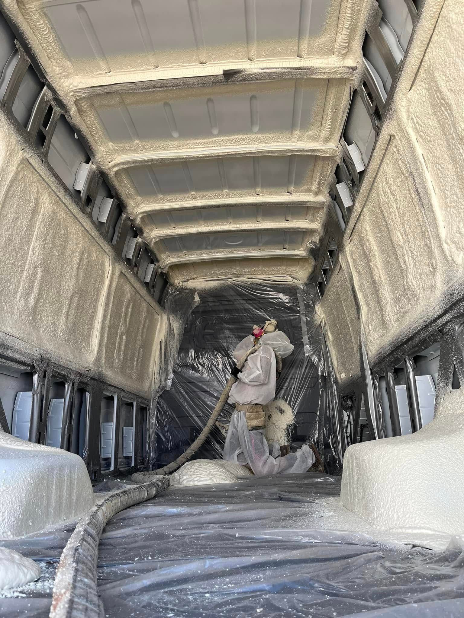 Person spraying insulation foam inside a van. Interior is covered in foam, with protective sheeting on the floor.