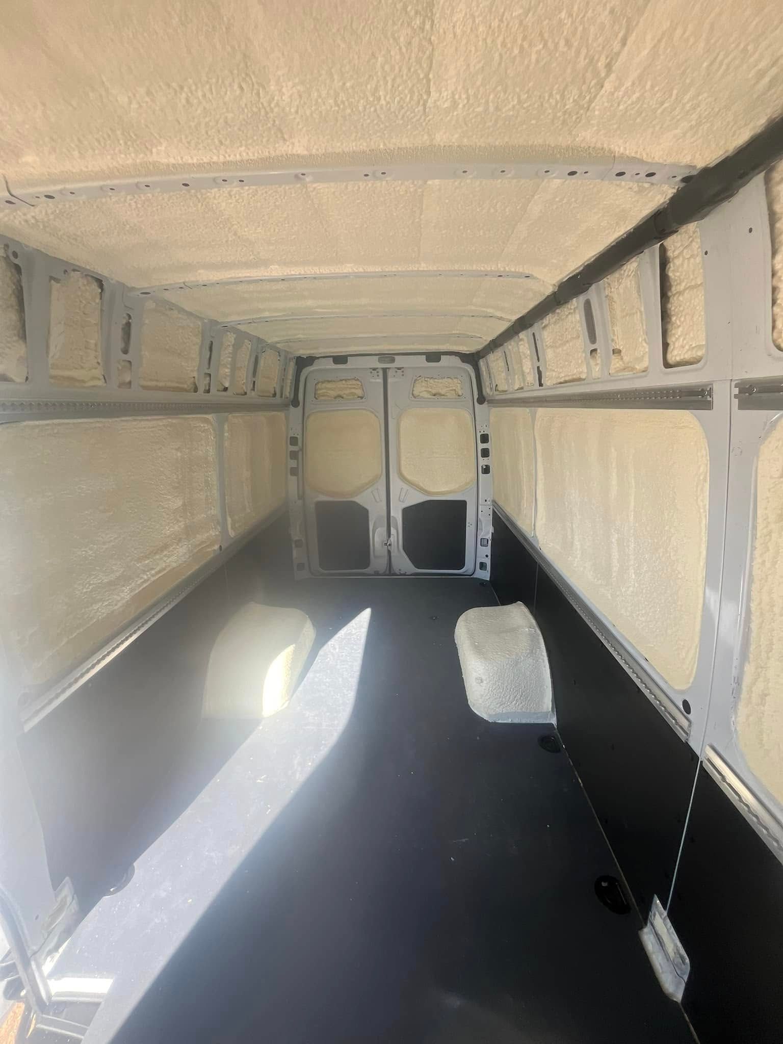 Interior of a van insulated with expanding foam, ready for conversion. Black floor, neutral-toned walls and ceiling.