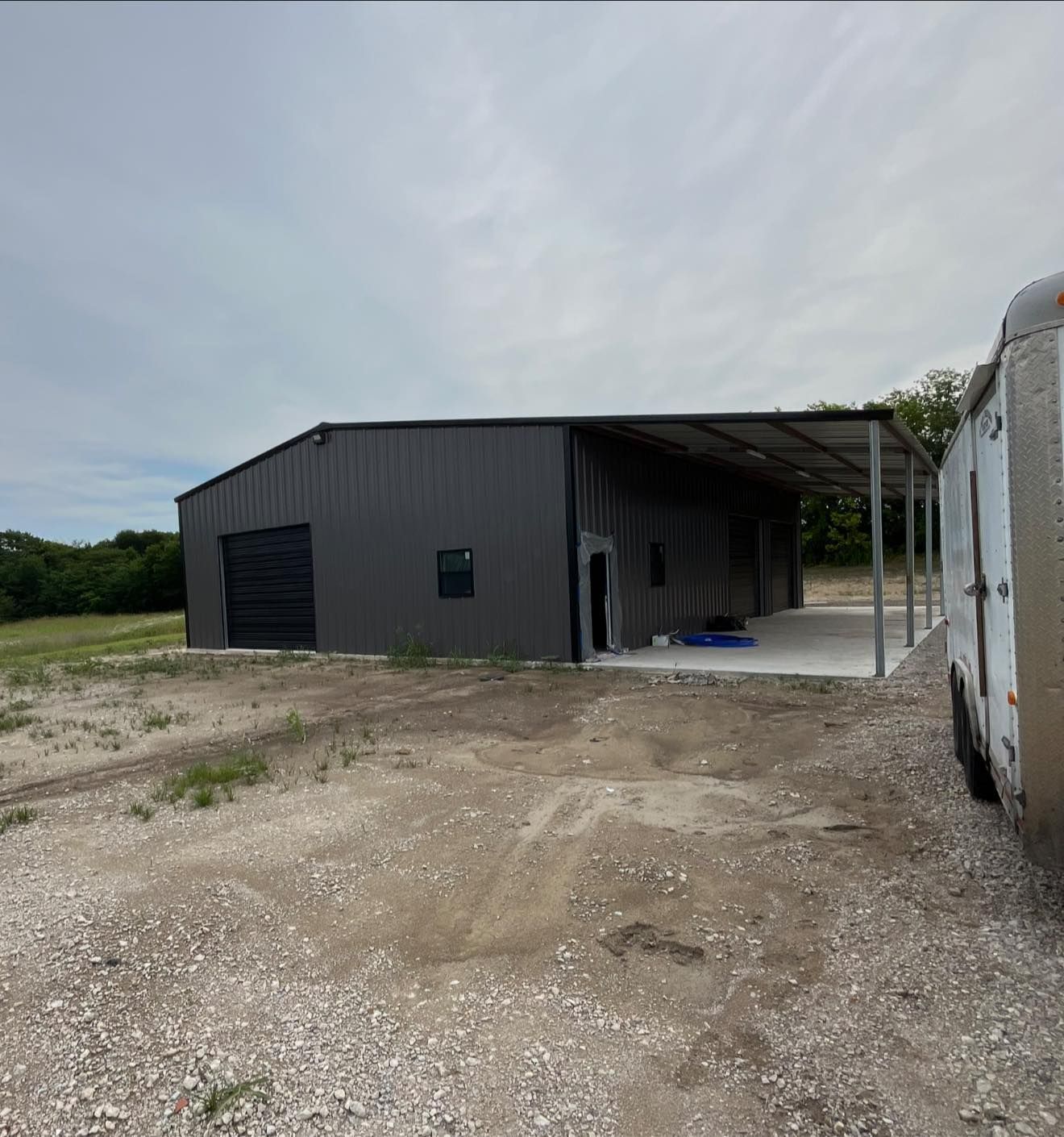 Gray metal building with covered porch and trailer on gravel.