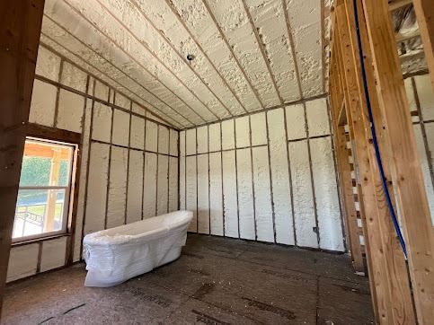 Interior room under construction, insulated with white spray foam. Wooden framing visible. Bag of insulation sits on the floor.