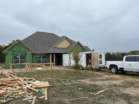 A house under construction with green wrap, a truck, trailer, and lumber on the ground.