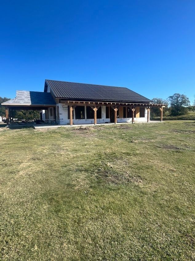 New house under construction with covered porch, wooden beams, and black roof, set on grassy field.