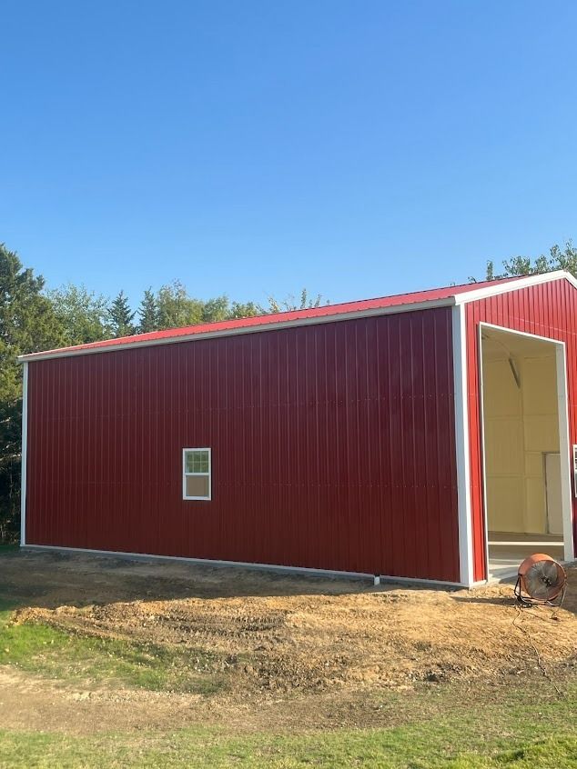 Red metal building with white trim, red roof, small window, and open doorway, set in a grassy area under a blue sky.