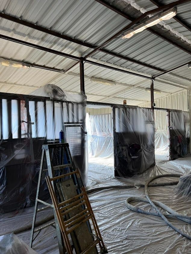 Interior of a building with plastic sheeting and spray foam insulation in progress. Ladder and hoses are visible.