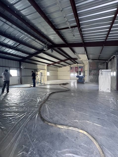 Workers insulating a large warehouse with spray foam. Plastic sheeting covers the floor.