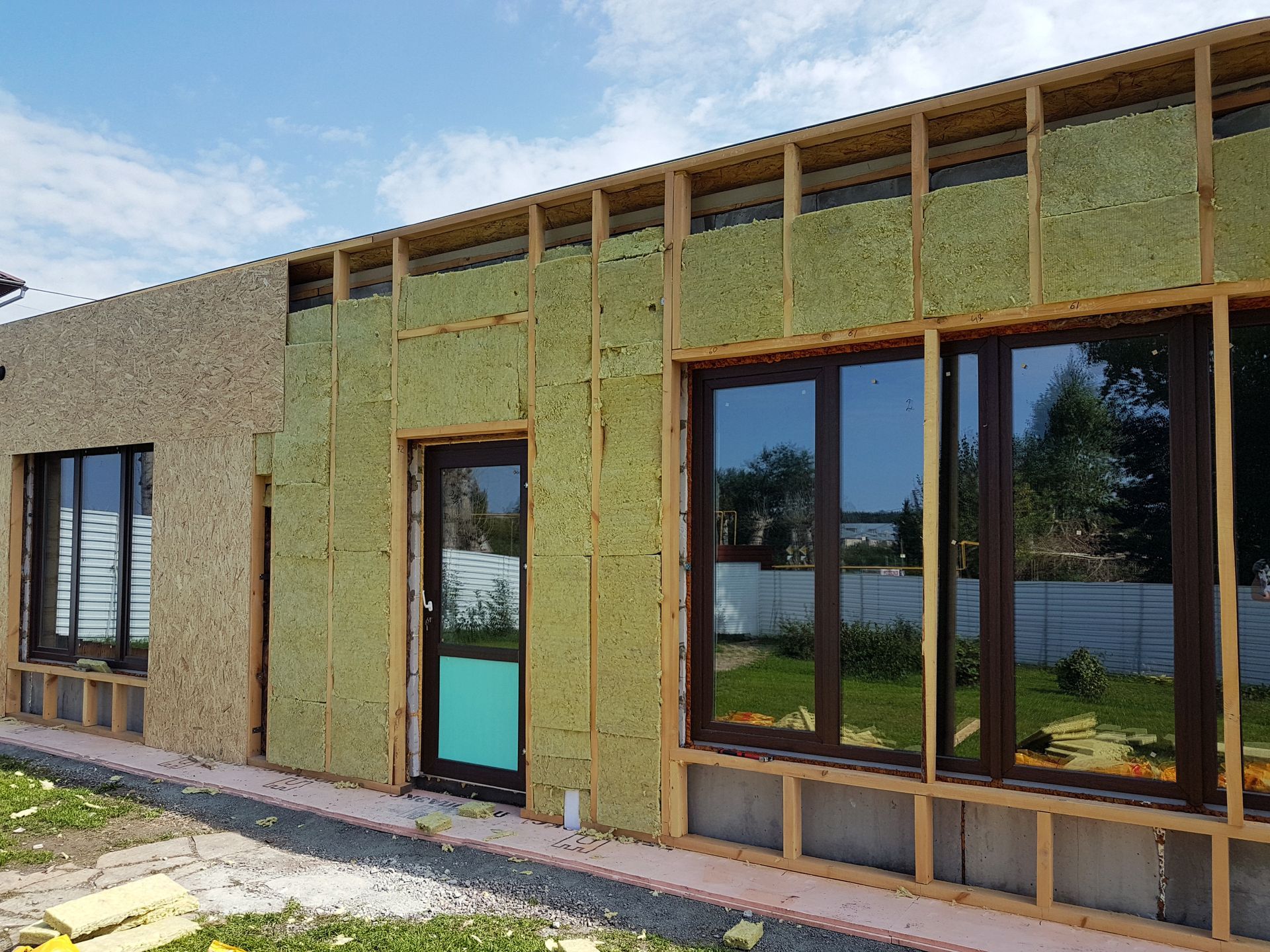 A house exterior under construction with exposed wooden framing, green insulation panels, and installed windows.