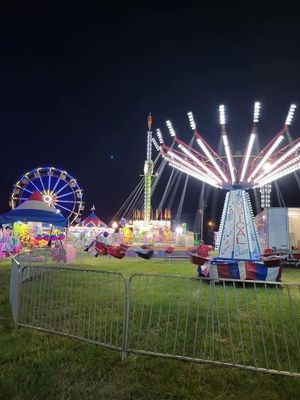 Nighttime fairground with a lit Ferris wheel, spinning rides, and colorful attractions behind metal barriers