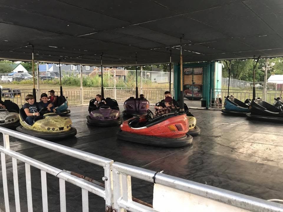 People riding bumper boats in a covered amusement park arena, with yellow, purple, orange, and black boats.