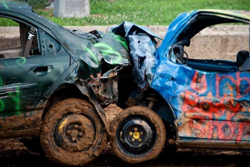 Two heavily damaged cars collided head-on, with crumpled green, blue, and red bodies and muddy wheels.